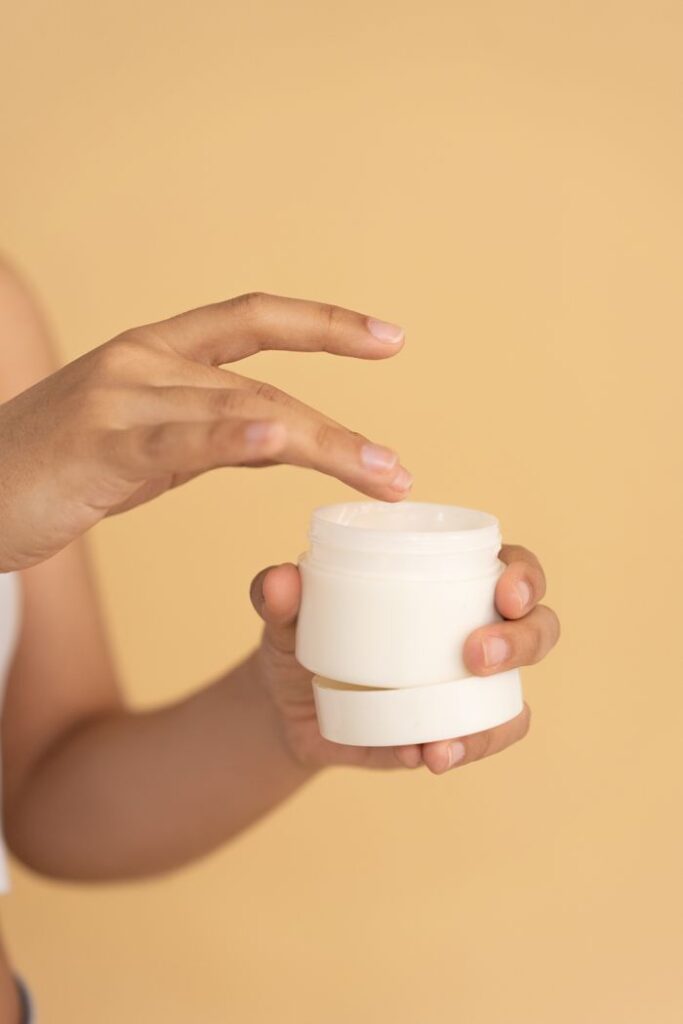 A woman's hand applying face cream from a jar against a beige background.